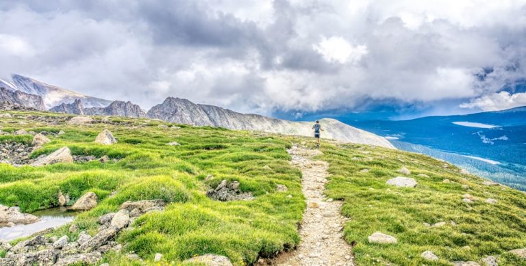 Two hikers with backpacks walking along a trail toward snow-capped mountains