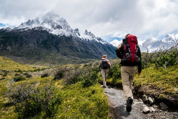 Two hikers with backpacks walking along a trail toward snow-capped mountains